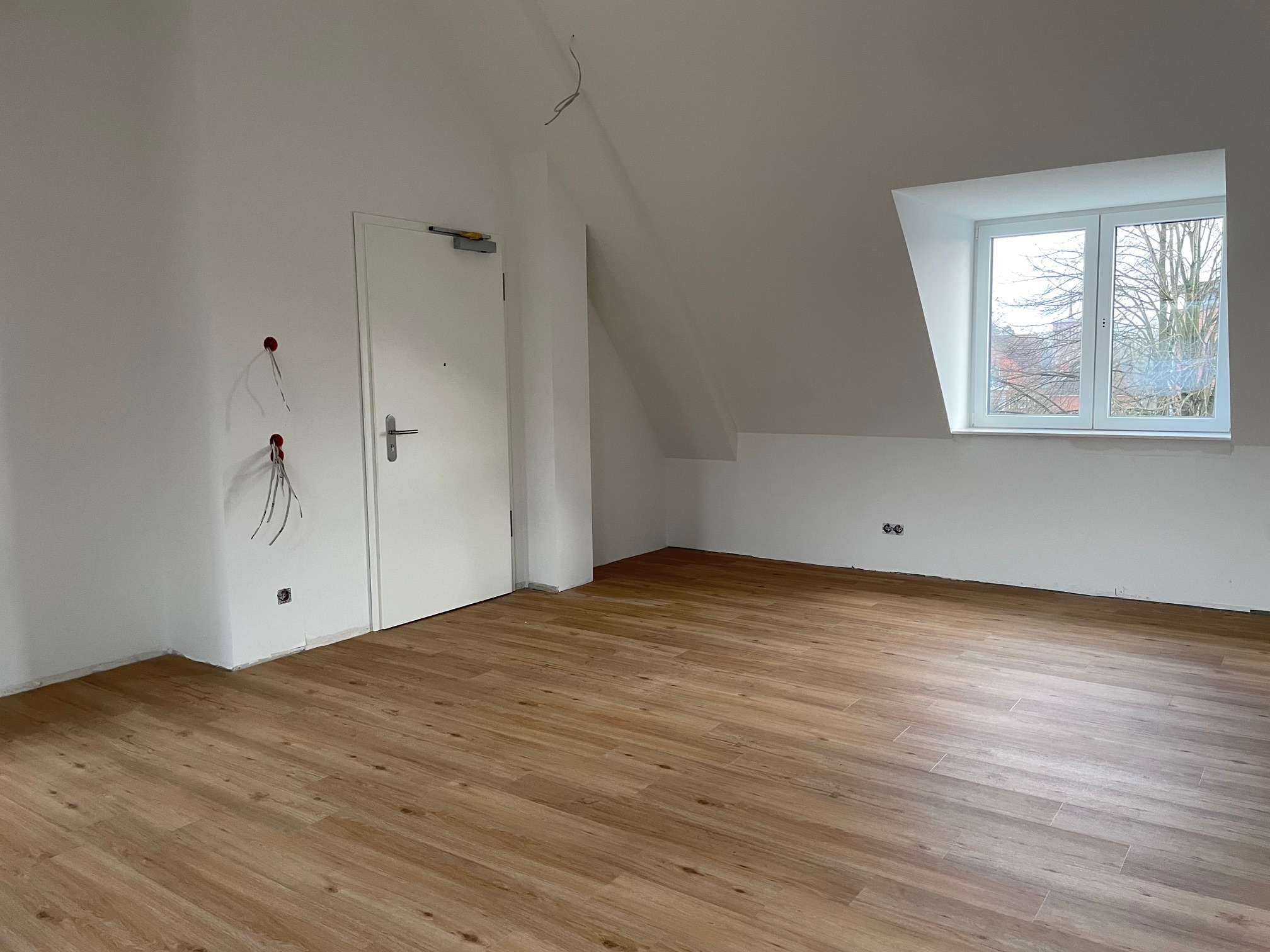 Leerer Raum mit Holzfußboden, weißer Tür, weiß gestrichenen Wänden und einem Fenster mit Blick nach draußen.